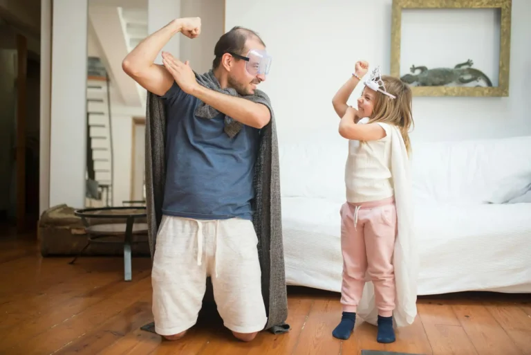 Nell'immagine, si vede un uomo che scherza con una bimba: entrambi hanno un braccio piegato per mostrare il muscolo. L'uomo indossa un paio di occhialoni trasparenti, la bimba ha una coroncina in testa.